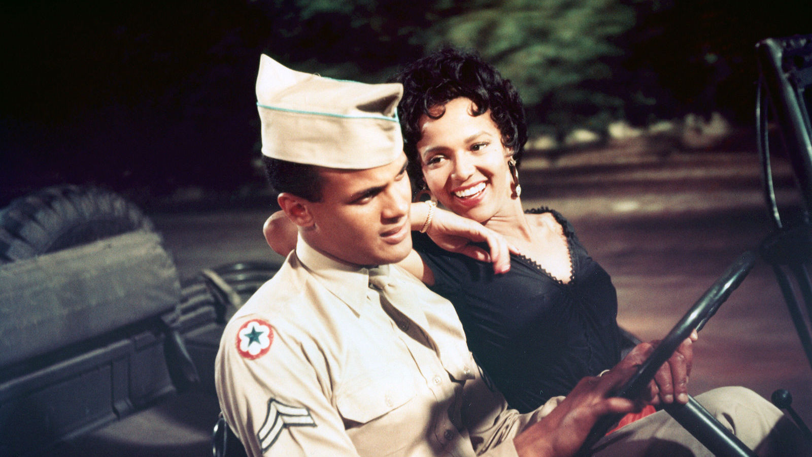 A man wears a military har and sits next to a woman in a car. She has her arm on his shoulder and smiles at him as he looks ahead.