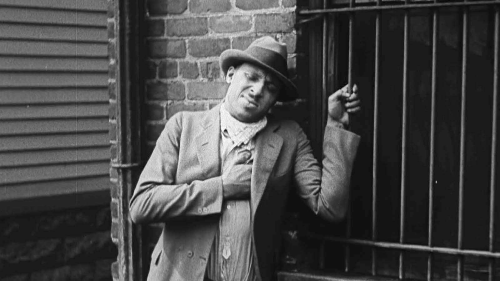 A man in a black and white photo stands agonized against the side of a brick city buliding, gripping iron window bars.