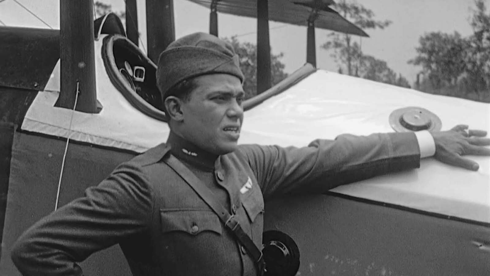 A fighter pilot in a black and white photo from early 20th century puts his hand on the side of a biplane.