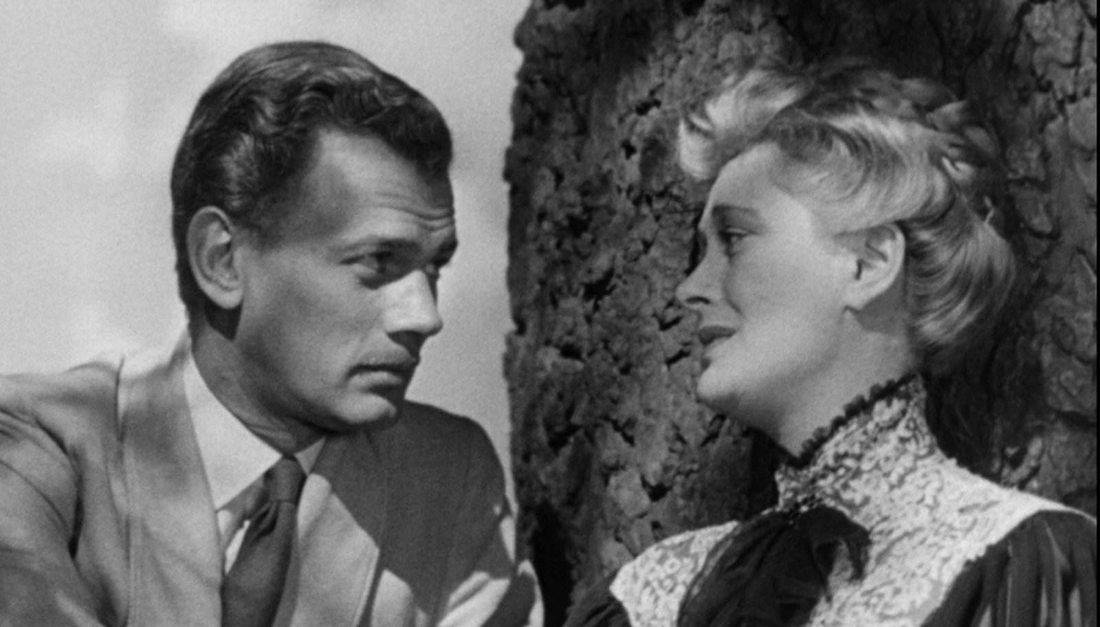 A mustachioed man looks longingly at a woman in a high starched collar leaning against a treek trunk