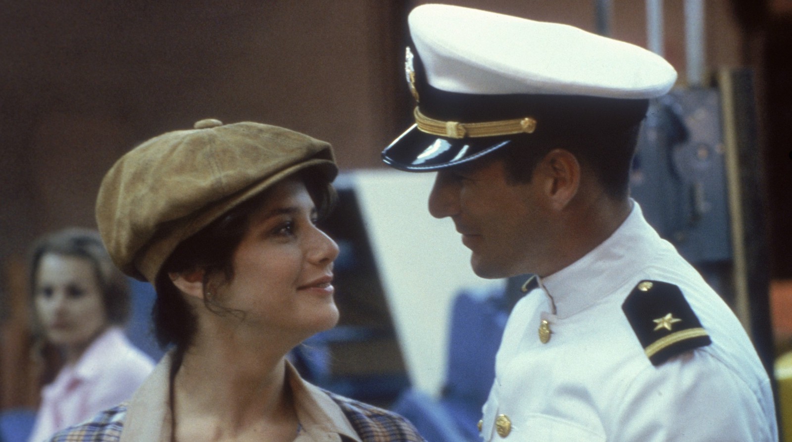 A woman in a pageboy hat and a man in a white cap with white naval officer's uniform stare at each other.