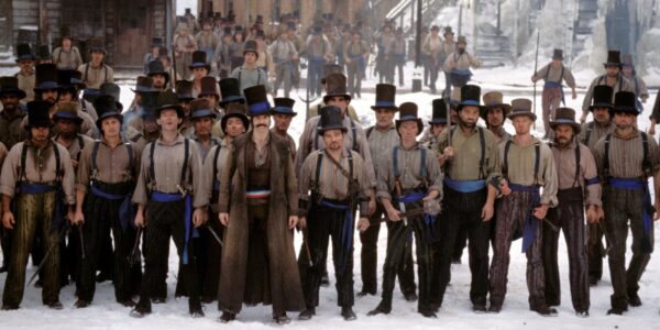 A line of tough-looking men in 18th century clothes, wielding weapons, are lined up in the snow with a threatening look.