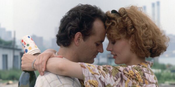 A dark haired man and a red headed woman embrace with their foreheads touching as they stand outside against a city skyline; she holds paper cups and champagne in her hand behind his back.