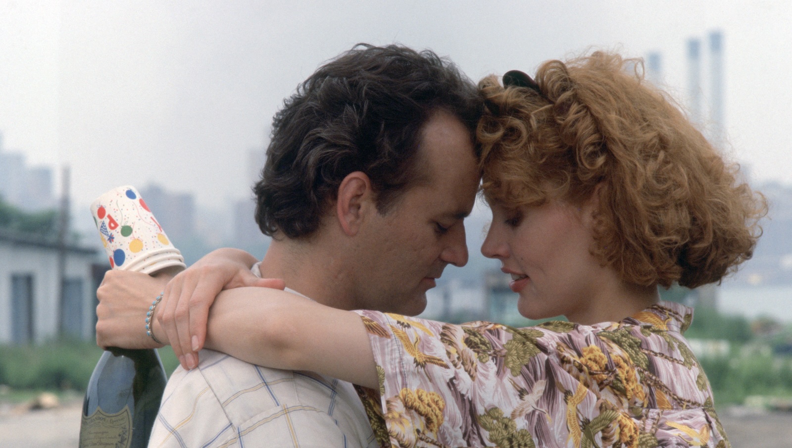 A dark haired man and a red headed woman embrace with their foreheads touching as they stand outside against a city skyline; she holds paper cups and champagne in her hand behind his back.