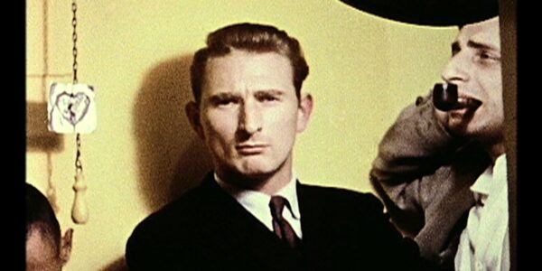 A man with coiffed hair and a suit and tie stares back from against a yellow wall.