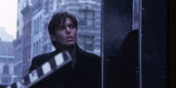 A man in a black coat stands outside in New York in front of a raised movie slate.