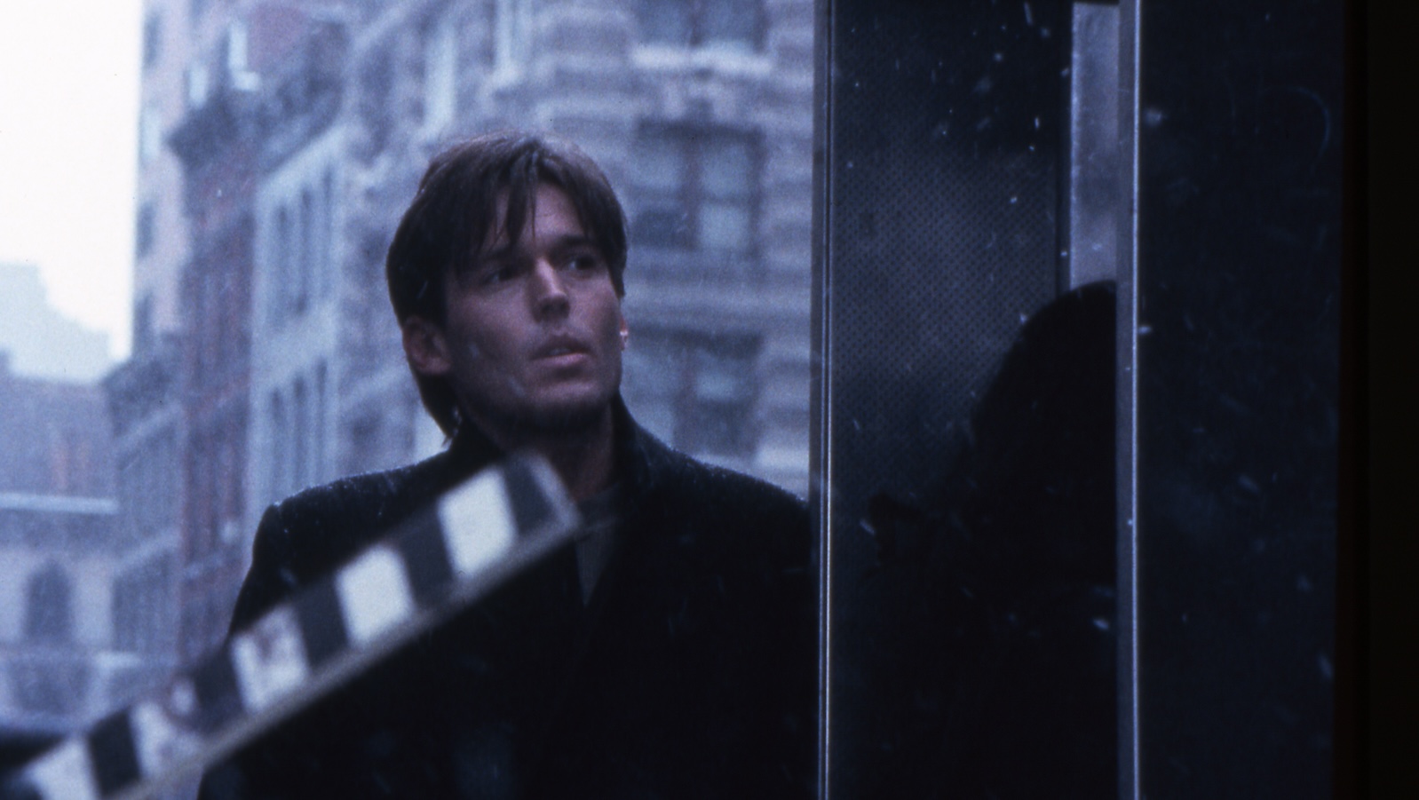 A man in a black coat stands outside in New York in front of a raised movie slate.