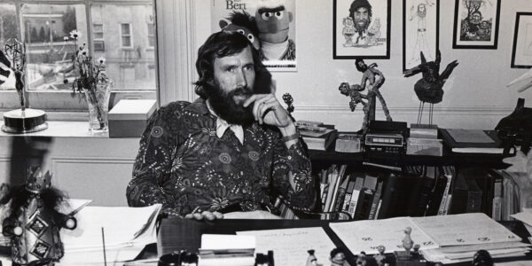 A bearded man sits at a desk talking on a phone is a black and white photo from the 1970s