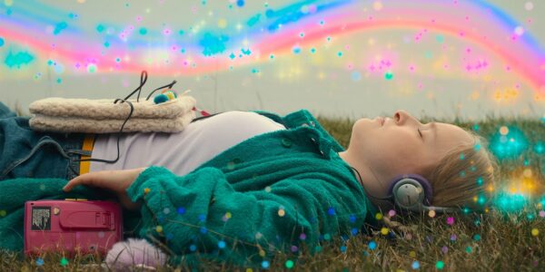 A young person lies on their back in a grassy field, eyes closed, wearing headphones and with a pink cassette player nearby. Colorful light effects and sparkles overlay the image.