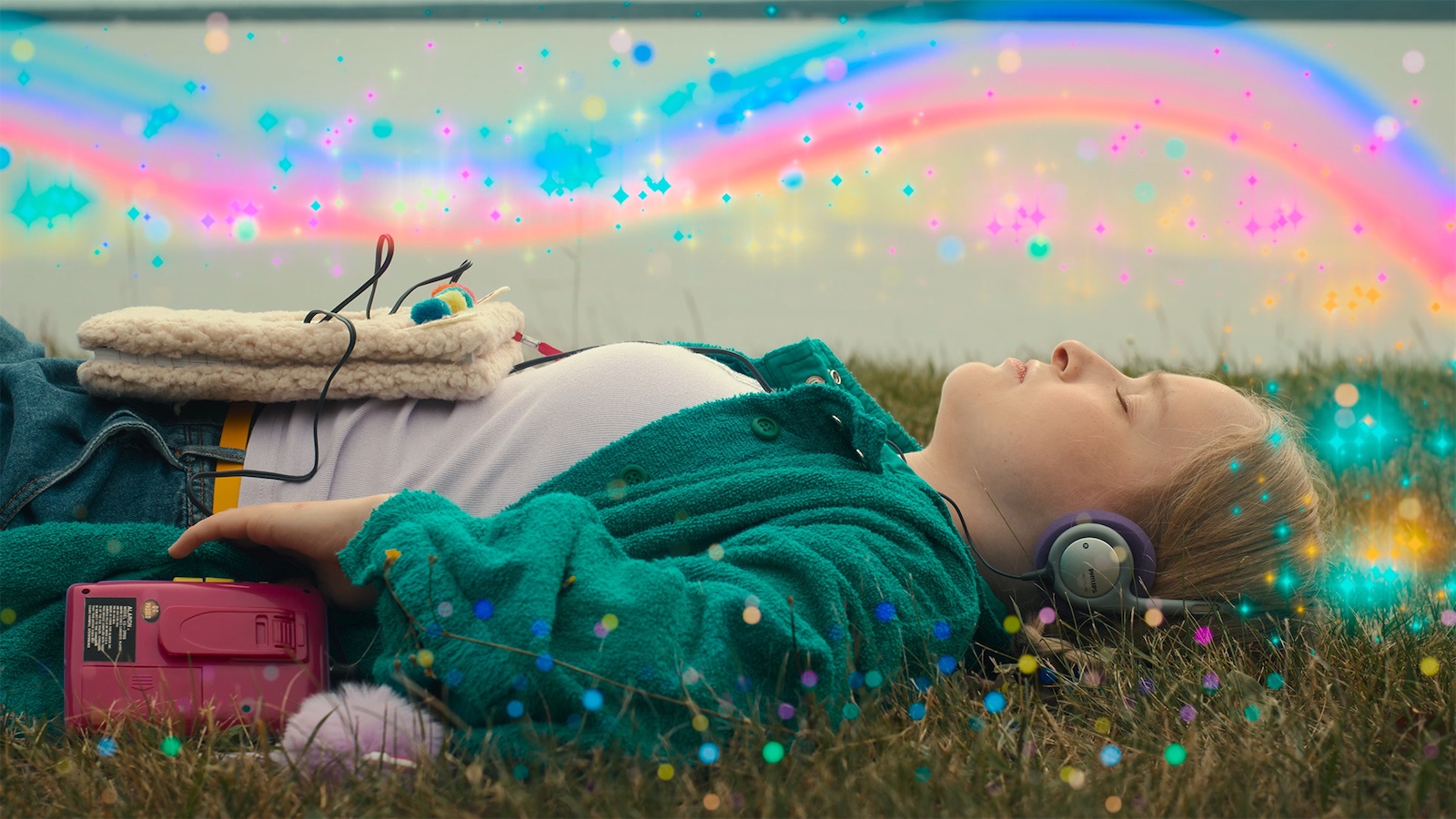 A young person lies on their back in a grassy field, eyes closed, wearing headphones and with a pink cassette player nearby. Colorful light effects and sparkles overlay the image.