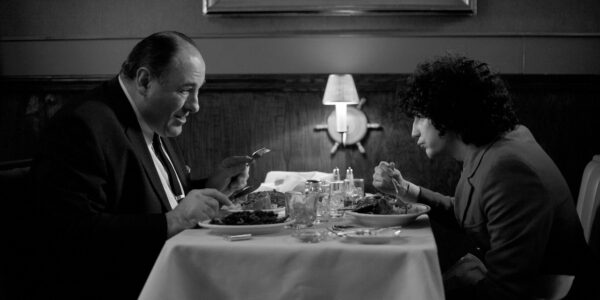 A middle-aged man and a teenage boy sit at a table in a restaurant, seen from profile, looking at each other intently as they eat, a single lamp between them.