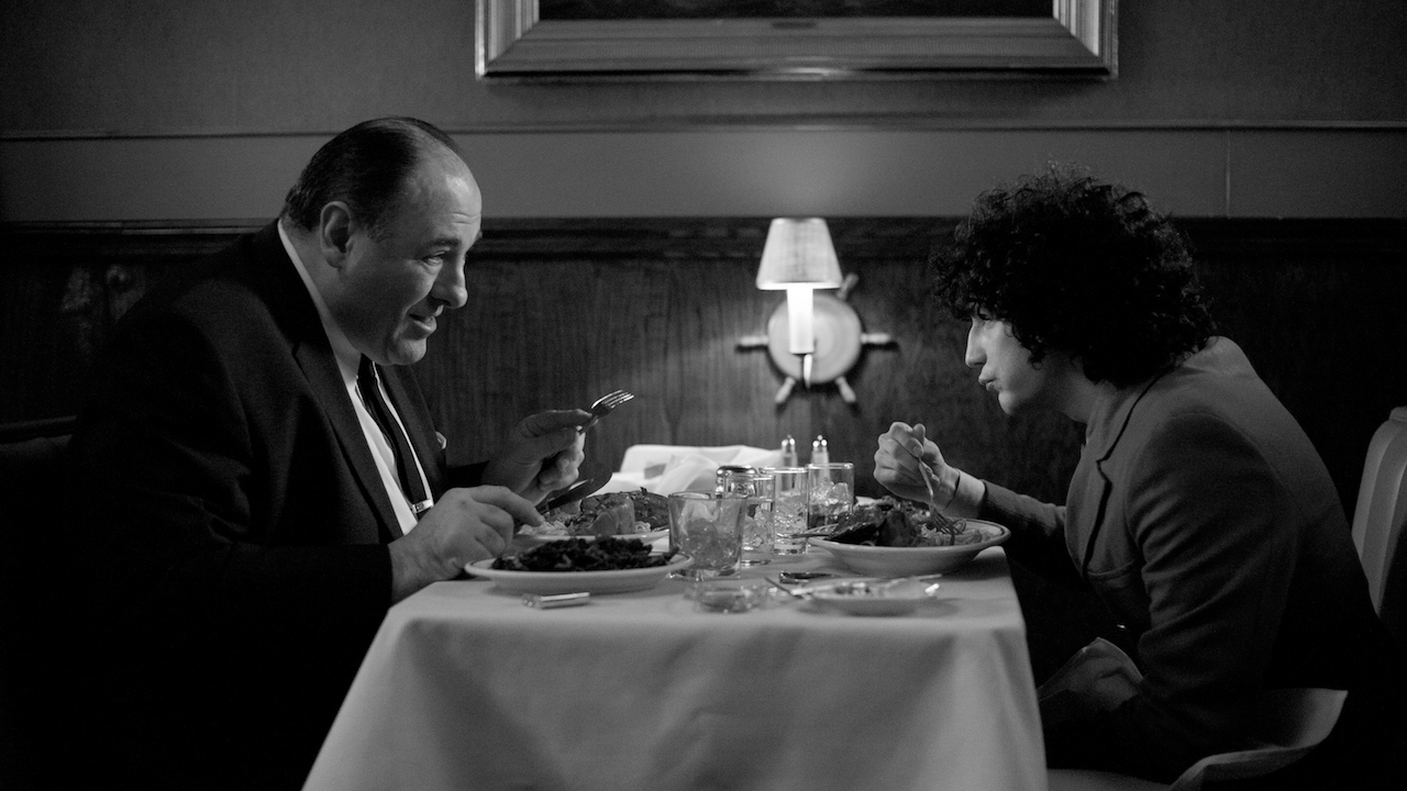 A middle-aged man and a teenage boy sit at a table in a restaurant, seen from profile, looking at each other intently as they eat, a single lamp between them.