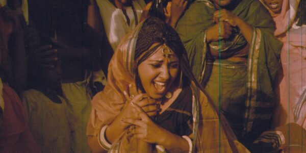 A woman in a sari screams with her mouth open, holding another person's hand as other women in saris stand around her.