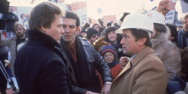 A man in a dark jacket looks intensely at a man in a beige coat and white plastic hard hat. Both are standing in profile amidst a political rally surrounded by placards with American flags, and a man in a leather jacket stands between them with an intense glare.
