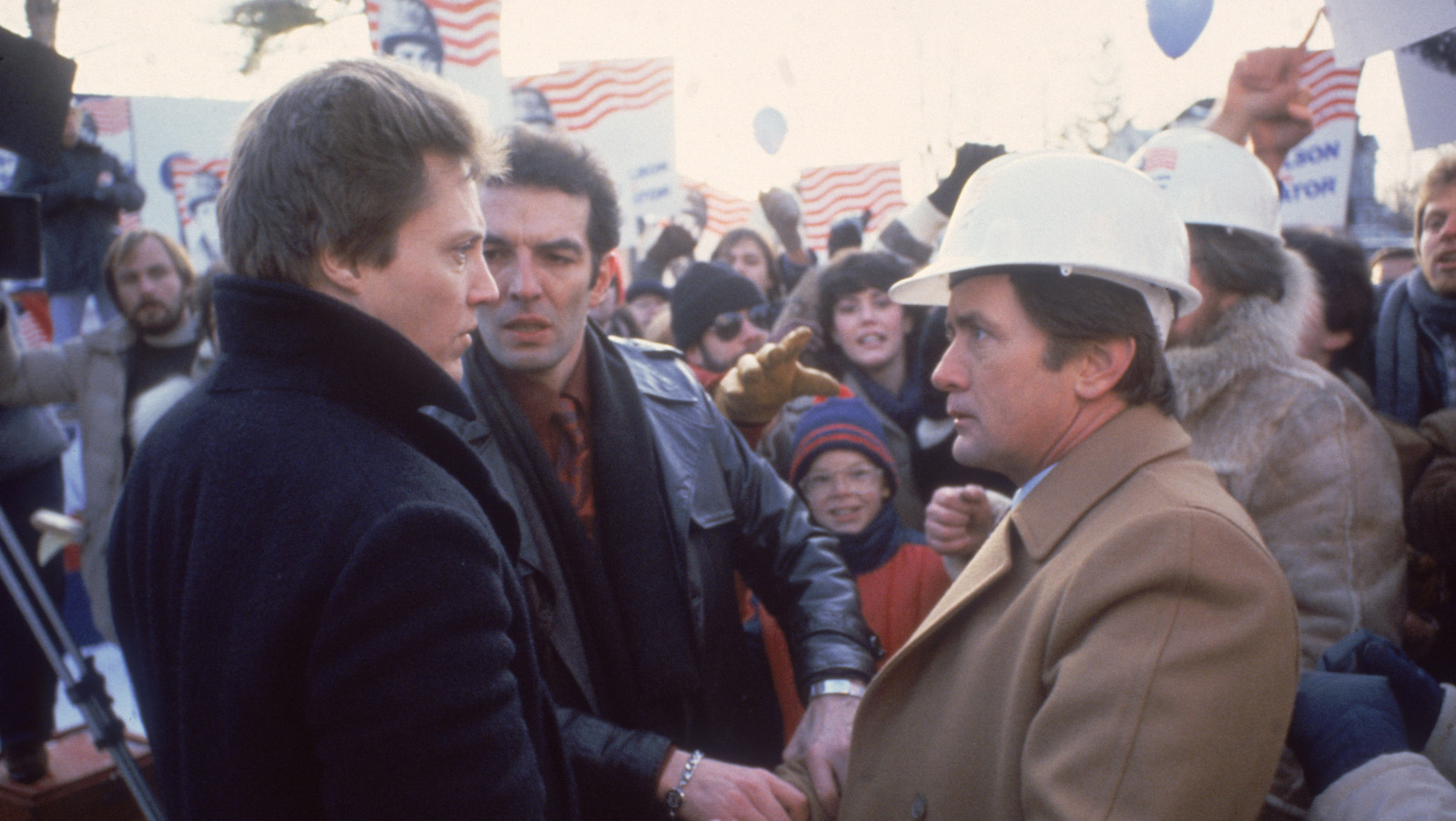 A man in a dark jacket looks intensely at a man in a beige coat and white plastic hard hat. Both are standing in profile amidst a political rally surrounded by placards with American flags, and a man in a leather jacket stands between them with an intense glare.