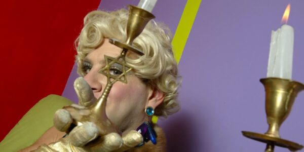 A gold-gloved drag performer in a blonde wig peers through a star of David in a candlestick.