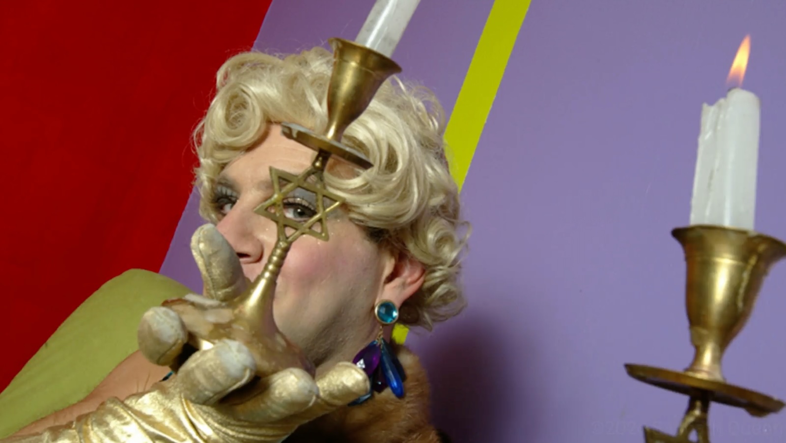 A gold-gloved drag performer in a blonde wig peers through a star of David in a candlestick.