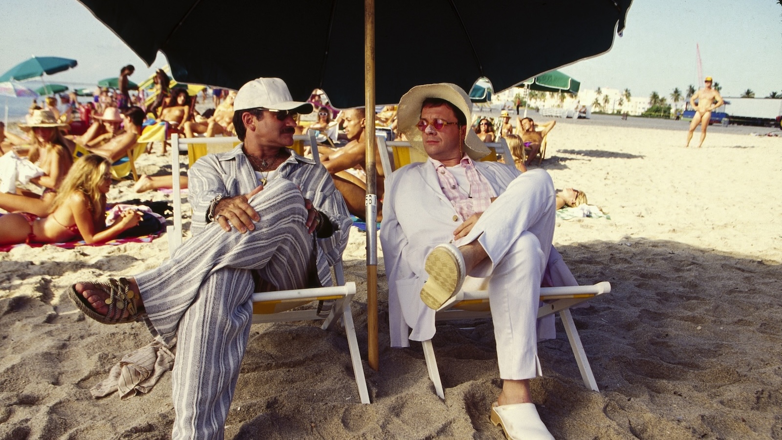 birdcage Two men sitting cross-legged on a beach, one wearing a sun hat, one with a ballcap, bothsitting under a large dark umbrella