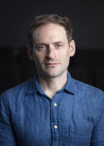 A serious looking man with short brown hair wearing a blue button-down shirt