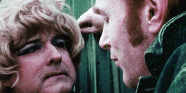 Grainy close-up of a man in a blond wig confronted by a man with an intimidating attitude in an old film style from the sixties.