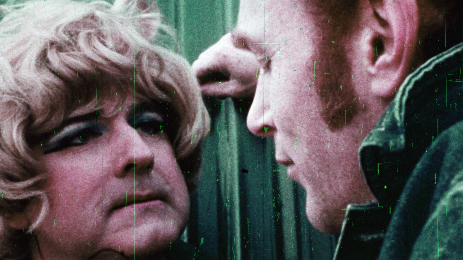 Grainy close-up of a man in a blond wig confronted by a man with an intimidating attitude in an old film style from the sixties.