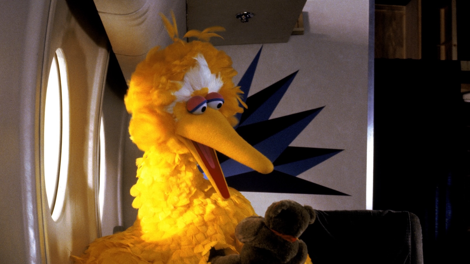 The big yellow muppet Big Bird sits on a plane next to a window holding a teddy bear. His beak is open wide and seems to be talking to it.