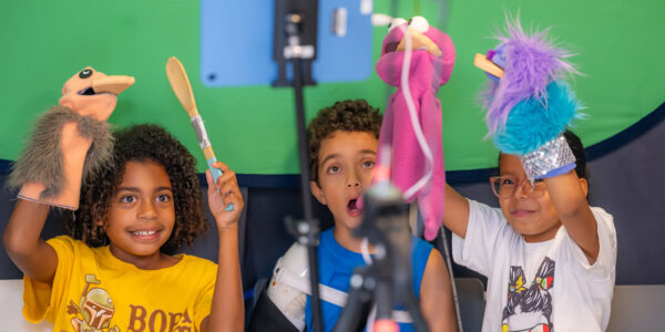 Three children hold up colorful puppets against a green screen; they are having fun, smiling and making funny faces with their mouths open, just like the puppets.