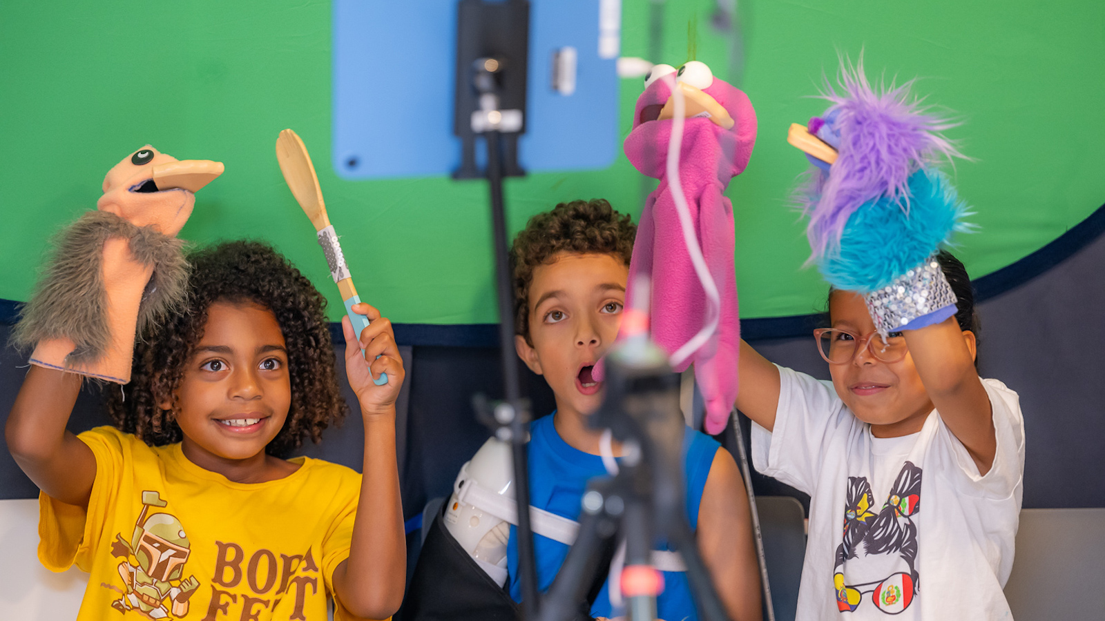 Three children hold up colorful puppets against a green screen; they are having fun, smiling and making funny faces with their mouths open, just like the puppets.