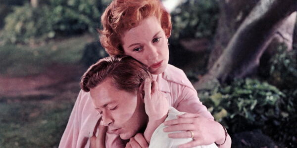 A red-headed woman embraces a dark haired man with tenderness as they crouch in a forest.