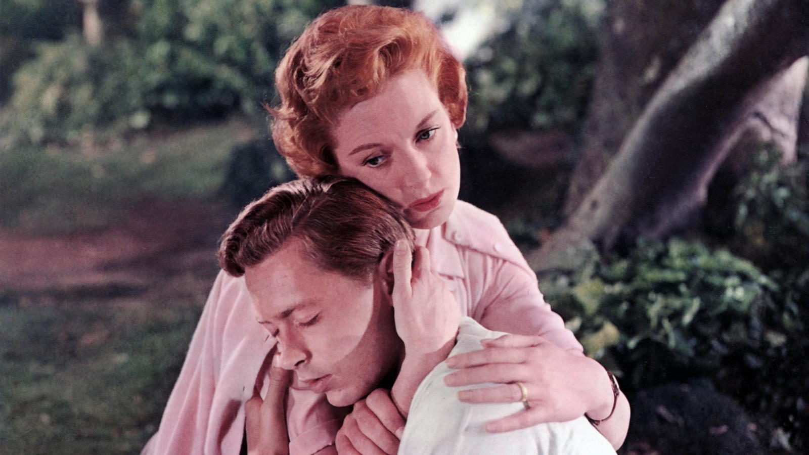A red-headed woman embraces a dark haired man with tenderness as they crouch in a forest.