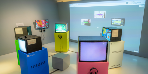 color photo of a museum installation showing 5 tube televisions on pedestals arranged like Stonehenge in a circle. Behind them on a wall is a large projection showing an internet browser.