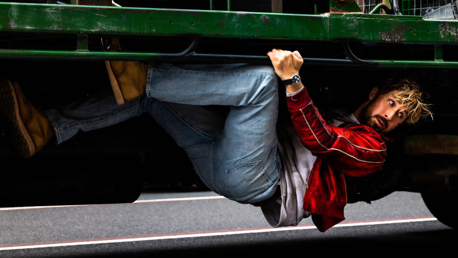A man in a red jacket and jeans dangles from the bottom of a truck.