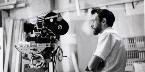 A man with a beard and coiffed hair and a white shirt with short sleeves looks off behind a film camera.