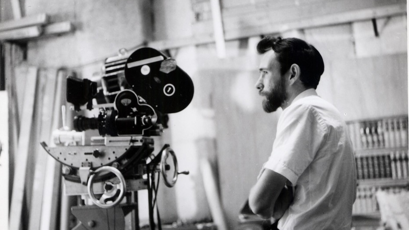 A man with a beard and coiffed hair and a white shirt with short sleeves looks off behind a film camera.
