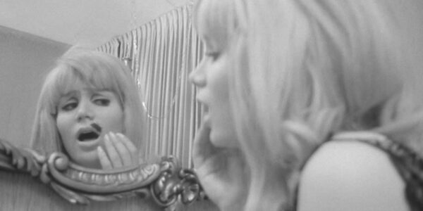 A blonde woman looks in a mirror, aghast by a black blotch on top of her lip.