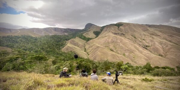 Against a vast mountain range, surrounded by fields of dry grass, a film crew of four crouches on the ground, facing a mountain, with a film camera and mic boom set up.