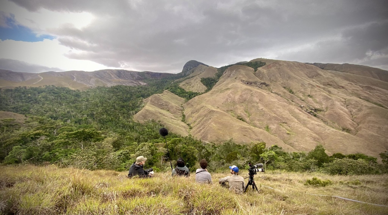 Against a vast mountain range, surrounded by fields of dry grass, a film crew of four crouches on the ground, facing a mountain, with a film camera and mic boom set up.